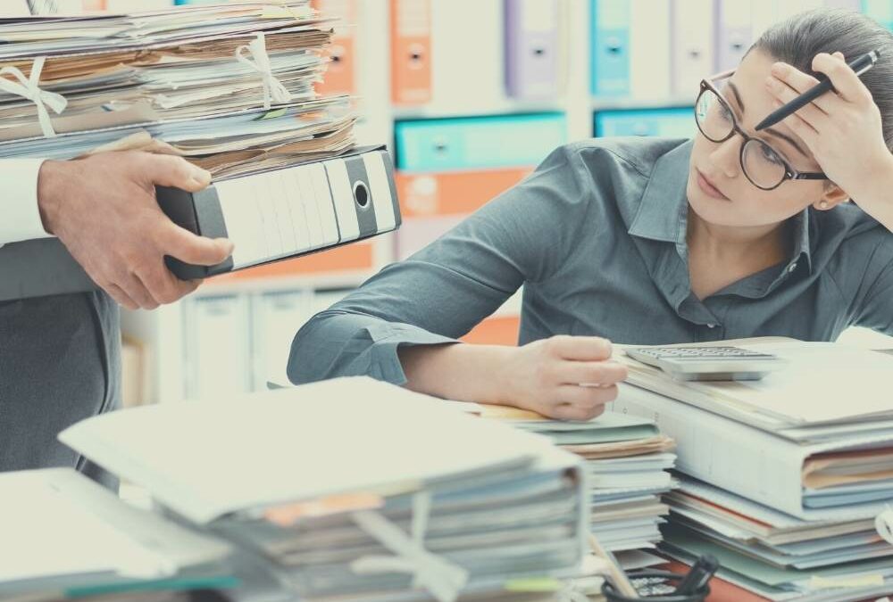 woman leaning her head into her hands, while sitting in front of piles of work, and more are comming.